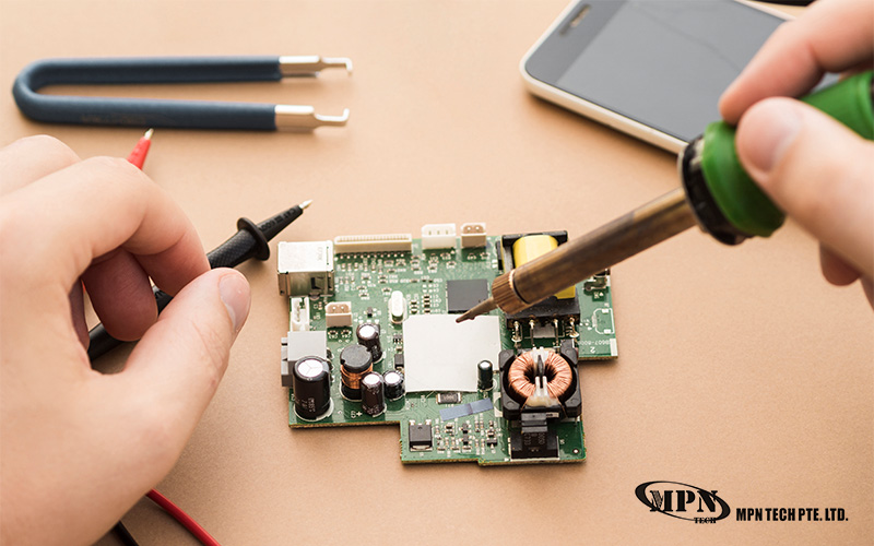 A person uses a soldering iron and multimeter probe on a green circuit board.