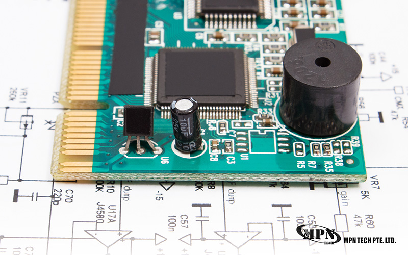 A green printed circuit board (PCB) sits on top of a white technical schematic diagram, showing various mounted electronic components like capacitors and microchips.