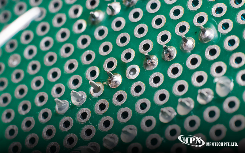 A macro close-up view of the underside of a green circuit board, highlighting silver solder points and metal component pins.